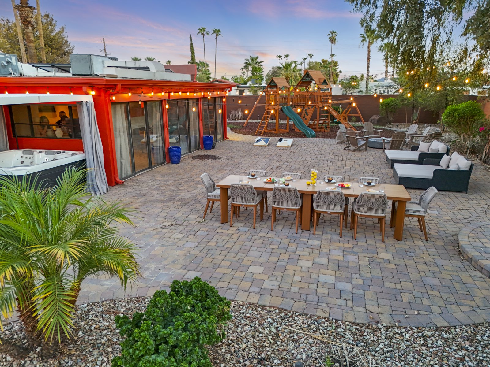A recent Scottsdale short-term rental shoot at twilight: string lights over a styled outdoor dining table, lit hot tub, playground and fire pit lounge under a pink desert sky.