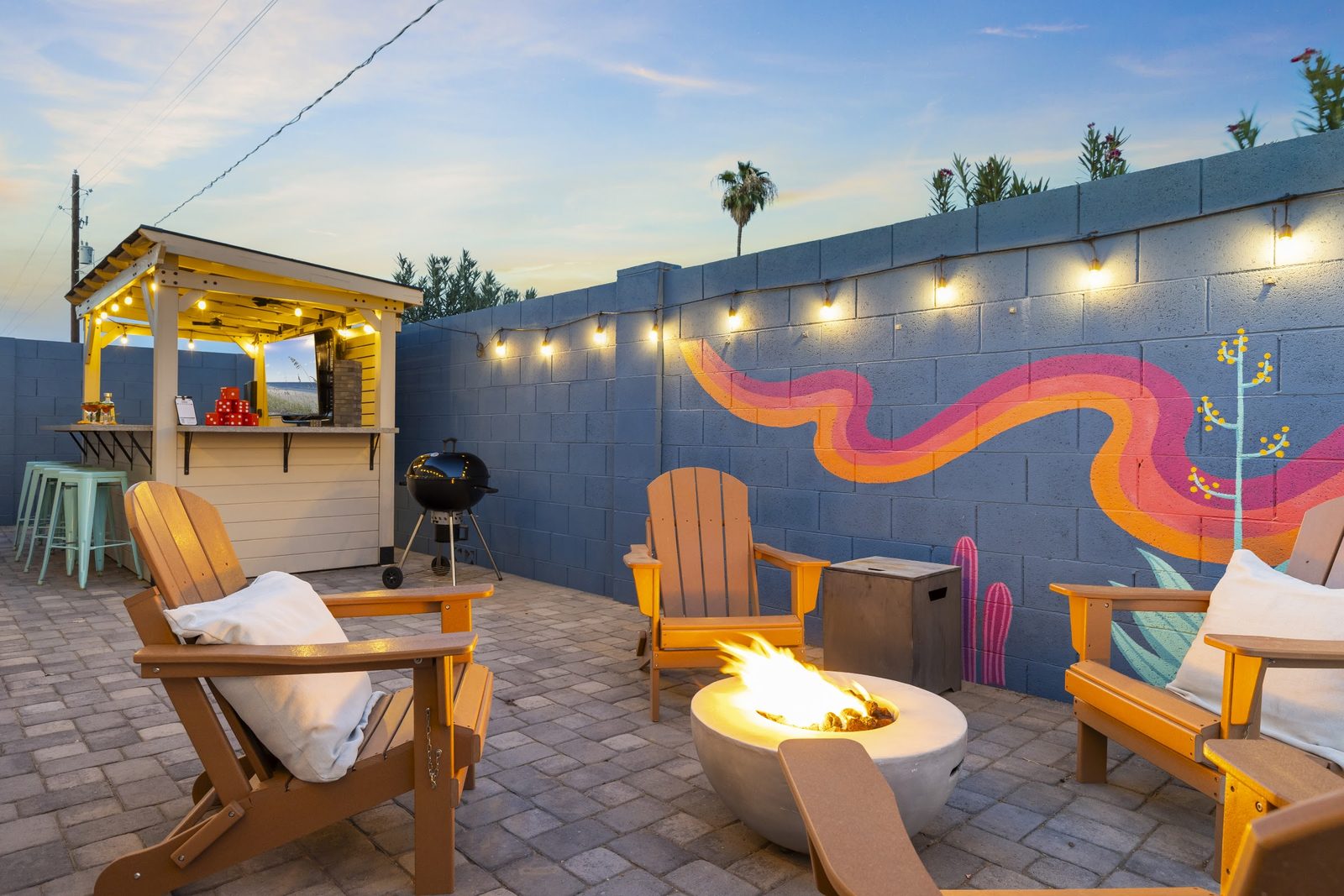 A styled fire pit scene at twilight: blankets over the chairs, drinks set out, fire lit, string lights on